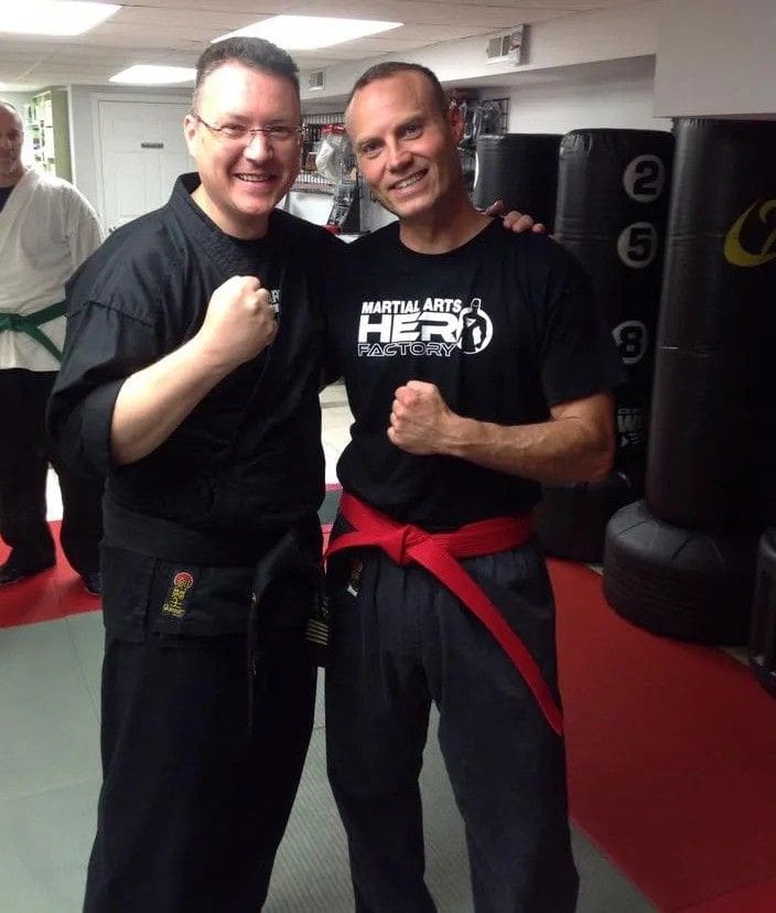 Two martial arts instructors wearing black uniforms and belts posing together inside a dojo.