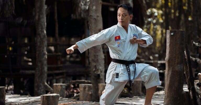 A martial arts progression timeline milestone showing an adult black belt instructor in a traditional white karatedo uniform, executing a sharp, disciplined side-punch stance outdoors on a rustic wooden platform.