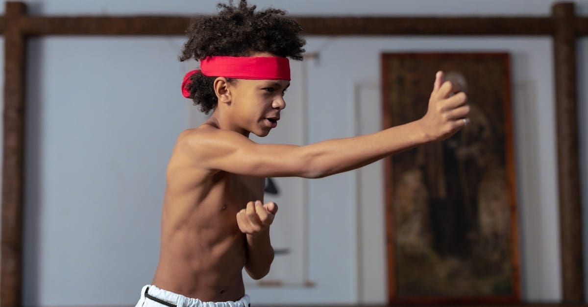 A side profile of a young boy with a red headband practicing a forward punch, capturing an early developmental stage in a martial arts progression timeline as he trains shirtless in a traditional dojo setting.