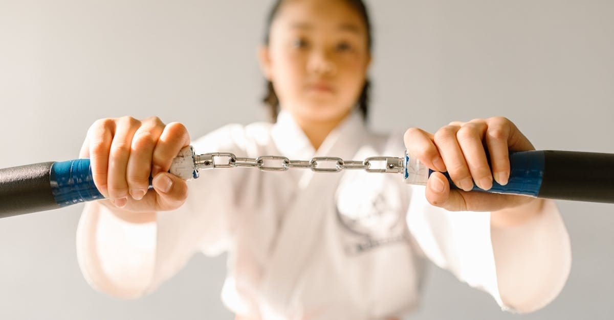 A close-up shot focusing on a young girl's hands tightly pulling a set of foam training nunchucks taut, marking the weapon-training phase of a martial arts progression timeline as she stands blurred in the background wearing a white gi.