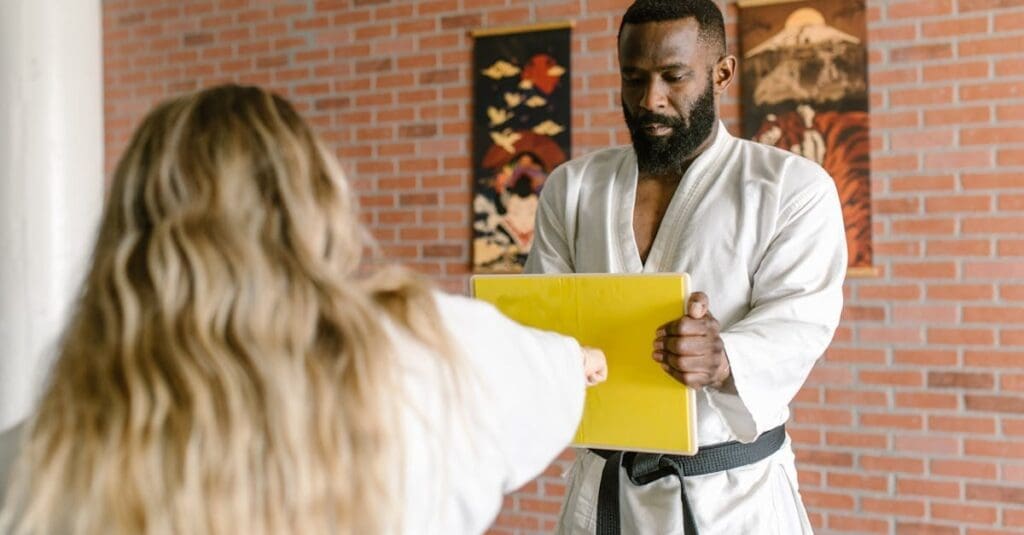 A side view of a bearded instructor in a white gi holding a yellow break-board for a student to strike, illustrating a common exercise in martial arts for beginners.