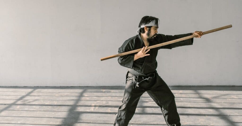 A martial artist in a black gi practices staff (bo staff) techniques, showing the variety of weapons training available in martial arts for beginners.