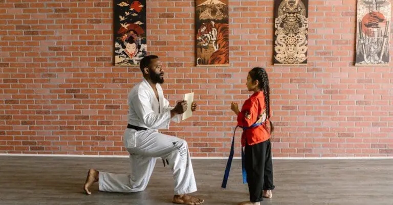 A bearded instructor in a white gi kneels on a wooden floor to guide a young girl in a red uniform through her first lesson in martial arts for beginners.